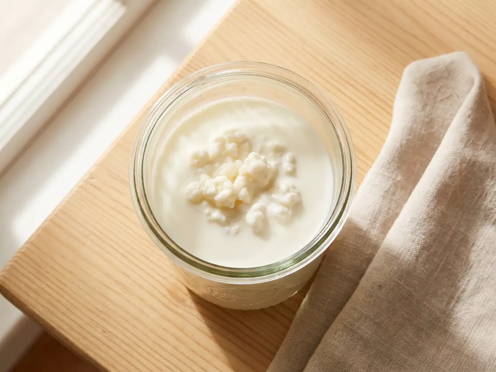 Tarro de vidro com kefir de leite a fermentar sobre madeira, junto a um pano de linho claro.