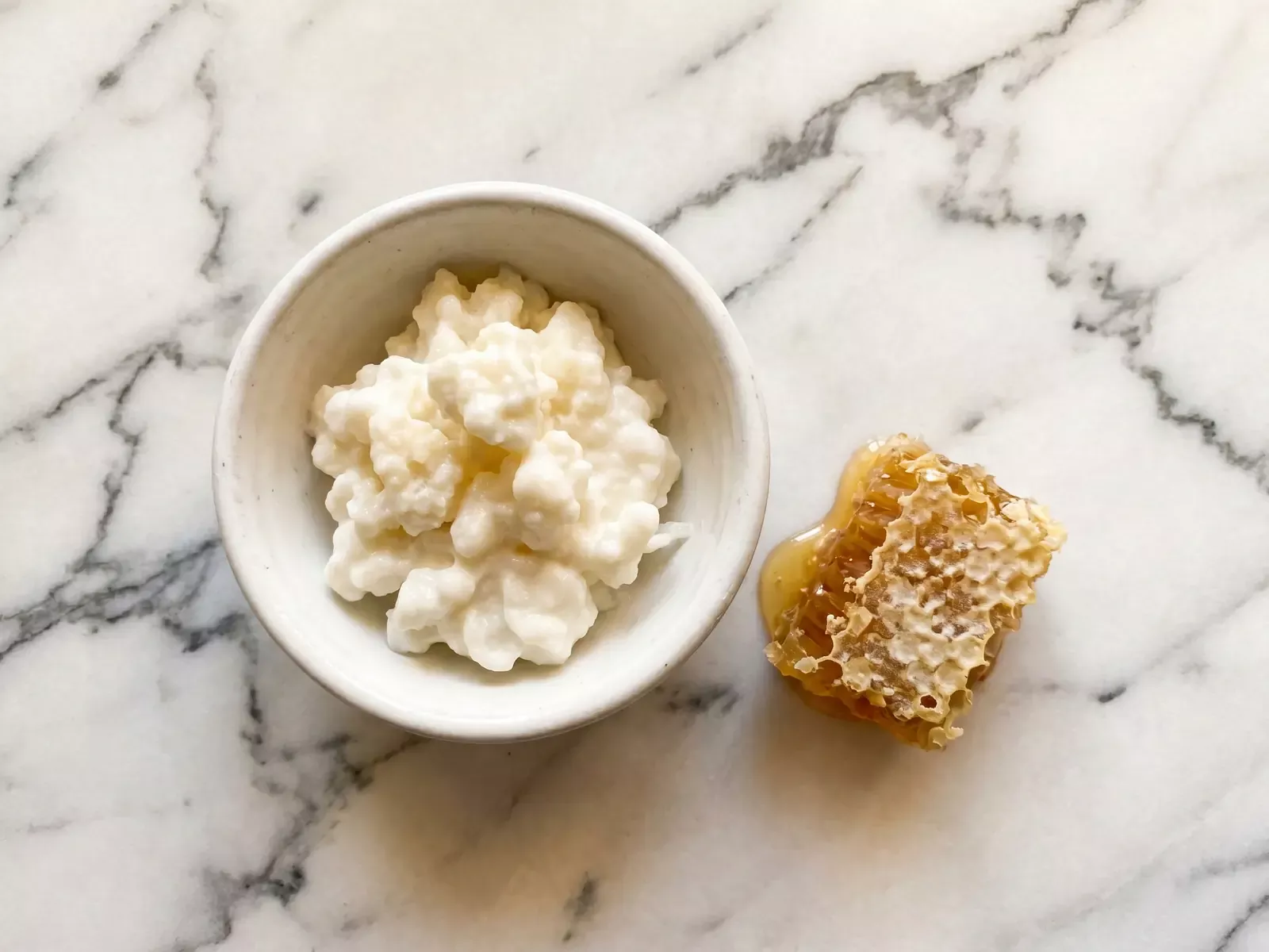 Cuenco de kefir natural com panal de mel ao lado, sobre m&aacute;rmore claro com luz cenital quente.